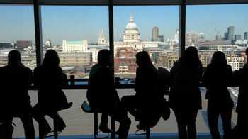 Tate modern silhouettes This is an urban landscape photograph taken inside the Tate Modern in London, United Kingdom, during the early afternoon in spring. The main subject of the image is the silhouettes of people sitting by the large window of the Tate Modern, with a clear view of London’s city architecture across the River Thames. The skyline is dominated by the prominent dome of St Paul’s Cathedral, a famous church and architectural landmark in London. The spring daylight provides a bright, clear backdrop, highlighting the city’s mixture of historic and modern buildings and emphasizing the iconic cathedral in contrast to the contemporary design of the Tate Modern.